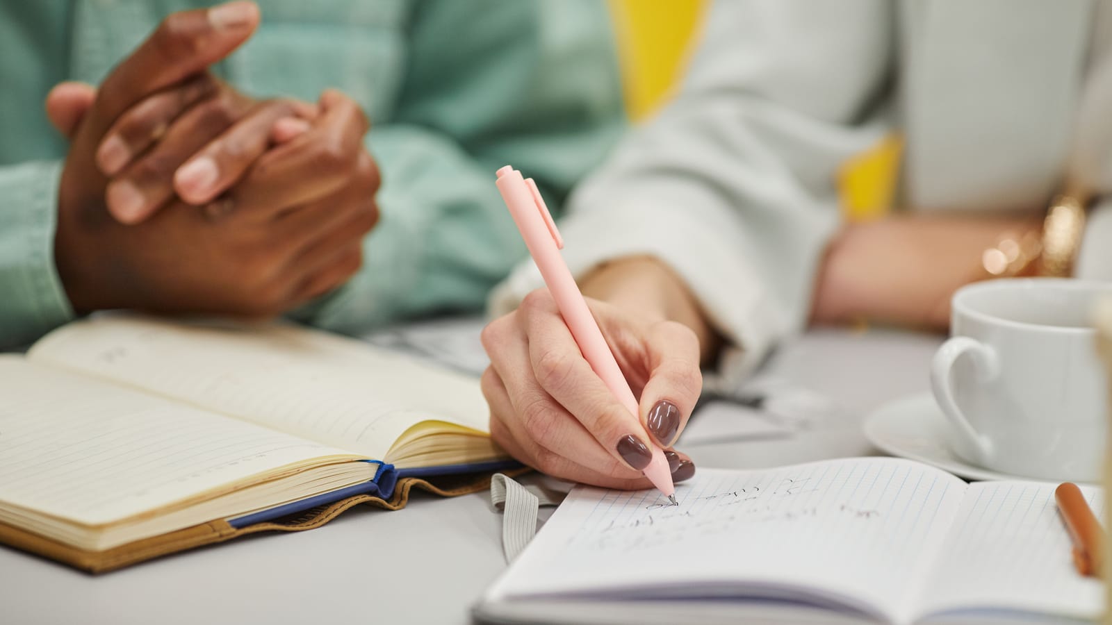 Toplantı masasında ciltli bir deftere kalemle yazı yazan el, yanında kenetlenmiş eller ve açık bir defter