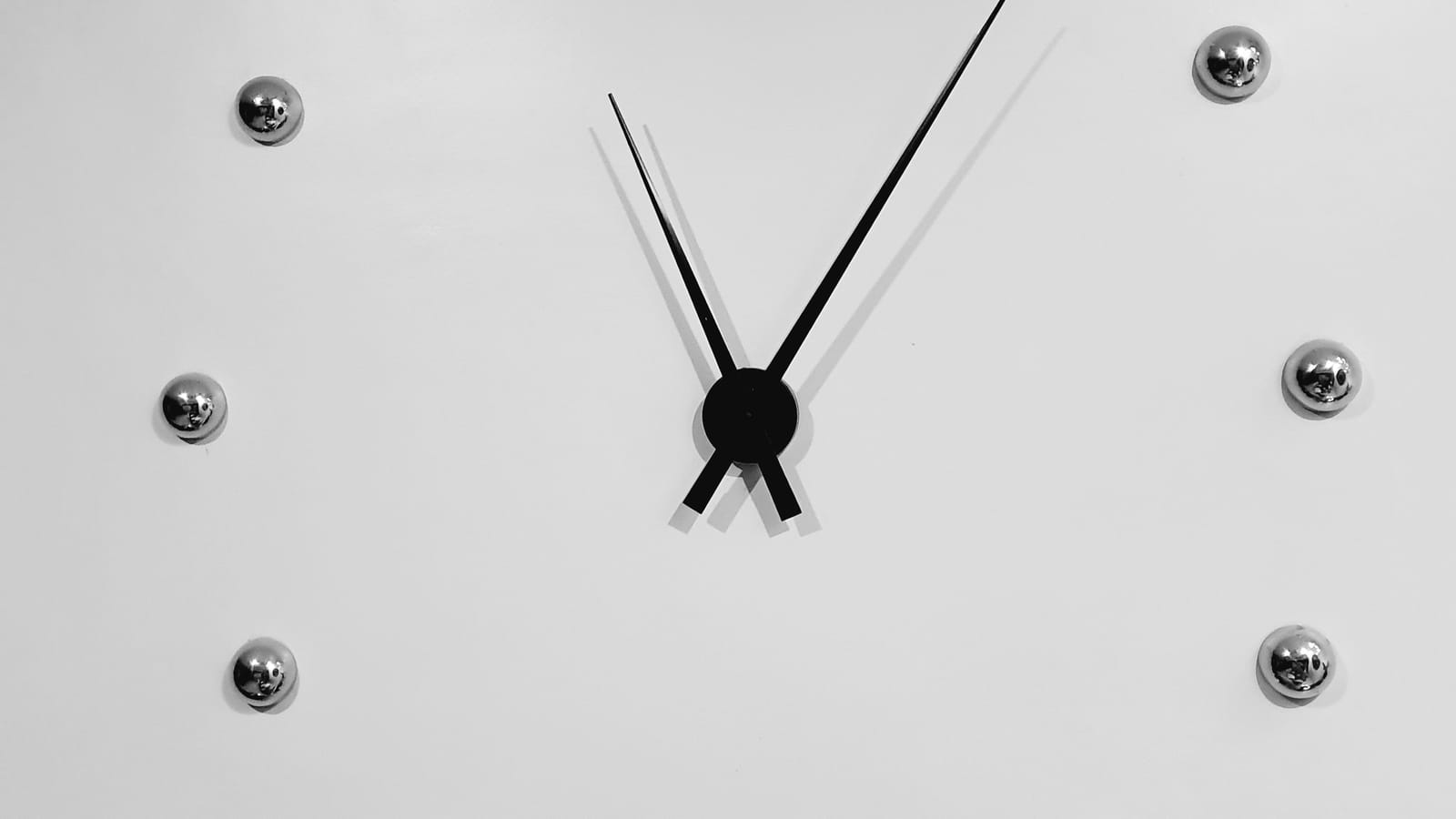 Beyaz duvarda rakamsız, yalnız metal noktalı minimal bir saatin akrep ve yelkovanı