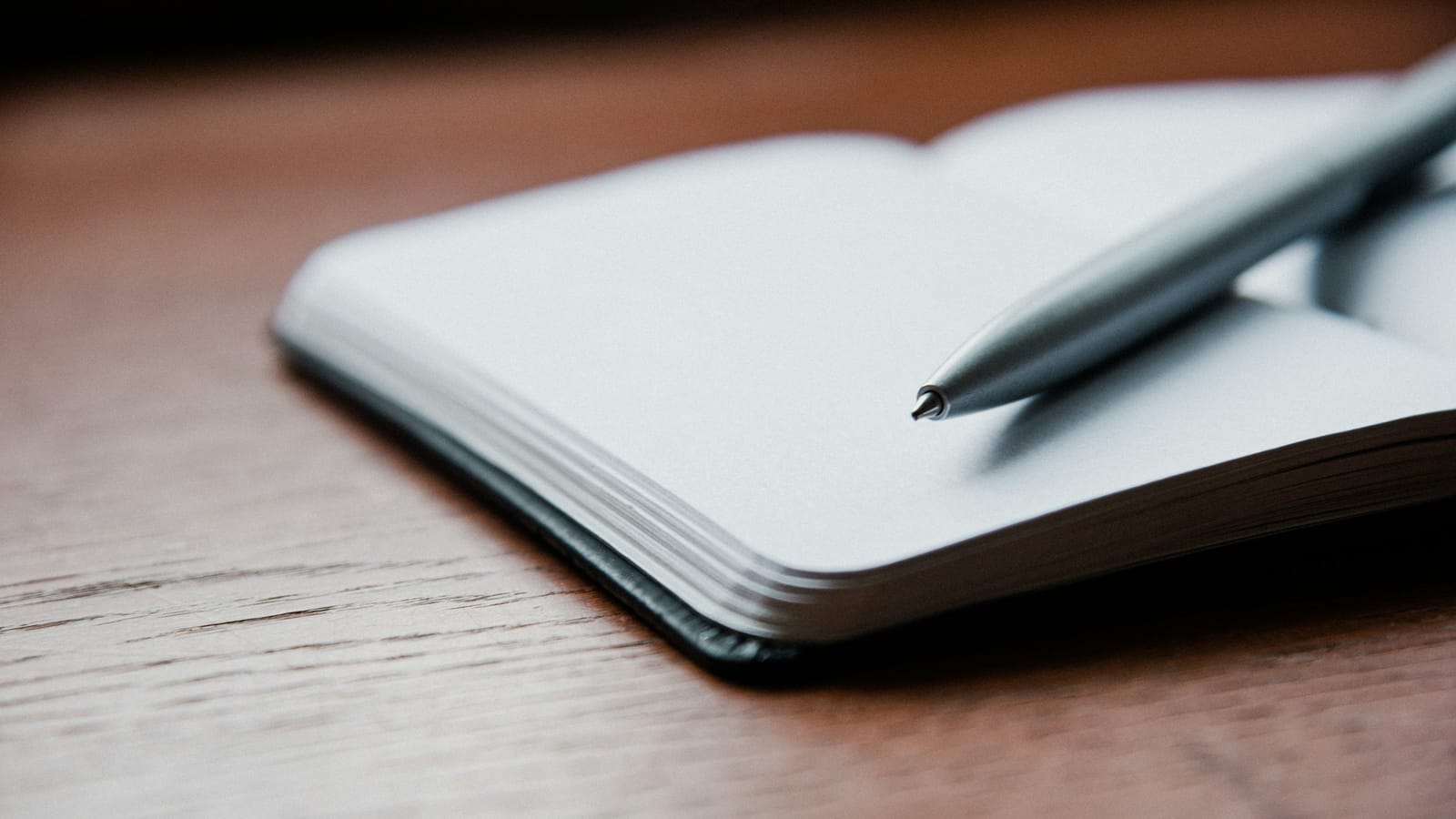 Ahşap masa üzerinde açık duran boş bir defter ve sayfaya uzanan gümüş renkli kalem