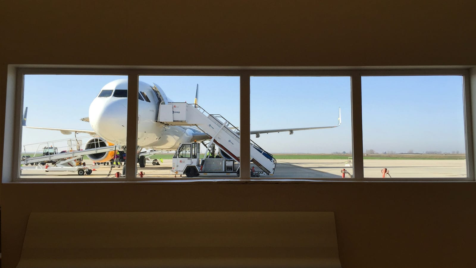 An aircraft parked at the gate, seen through an airport terminal window above an empty bench