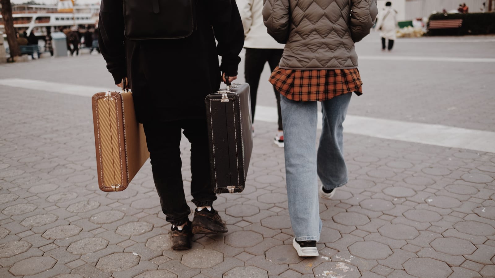 Two travellers seen from behind carrying vintage suitcases across a paved waterfront square in Istanbul